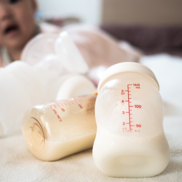 Duas mamadeiras transparentes cruzadas com leite e ao fundo a imagem ofuscada de um bebê.